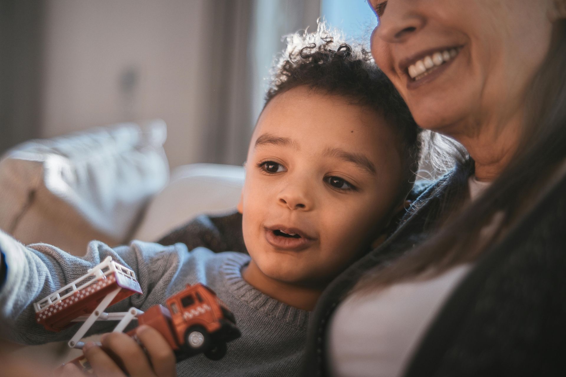 Boy holding toy fire truck, looking up with excited expression, sitting with smiling person.