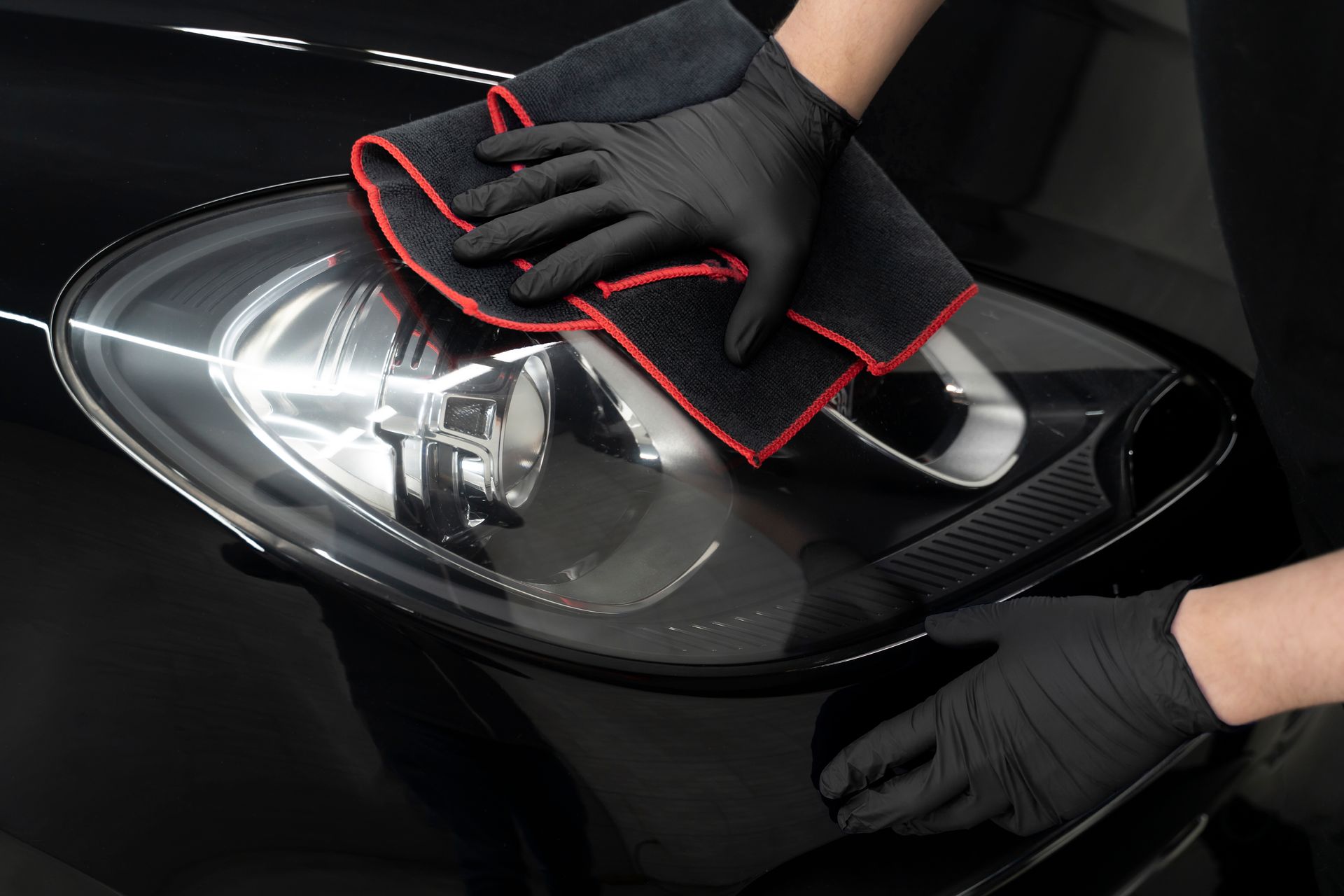 A person wearing black gloves is cleaning the headlight of a black car.