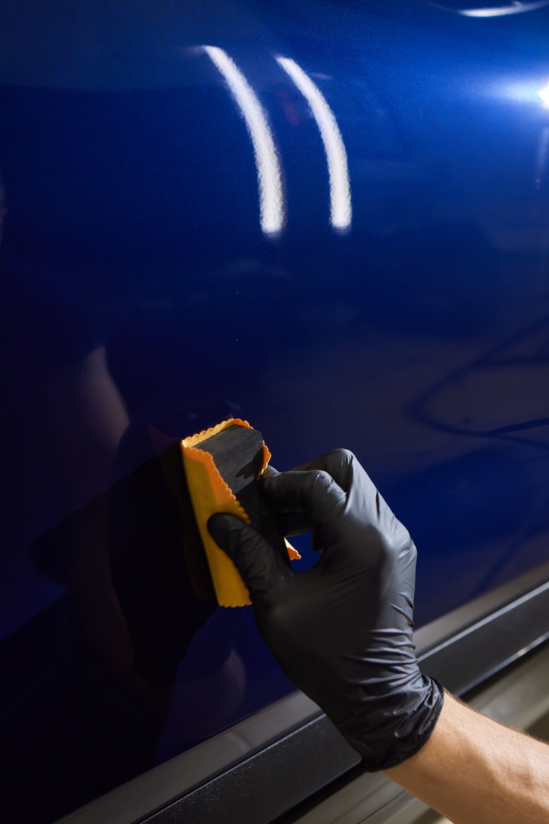 A person wearing black gloves is polishing a blue car with a yellow sponge.