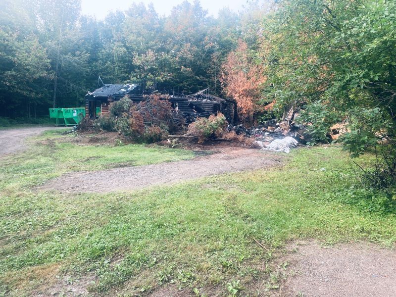A charred structure surrounded by vegetation and a dirt path. Green grass and trees in the background.