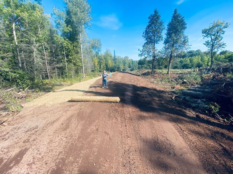 Person unrolling sod on a wide dirt road under a bright blue sky, surrounded by trees.