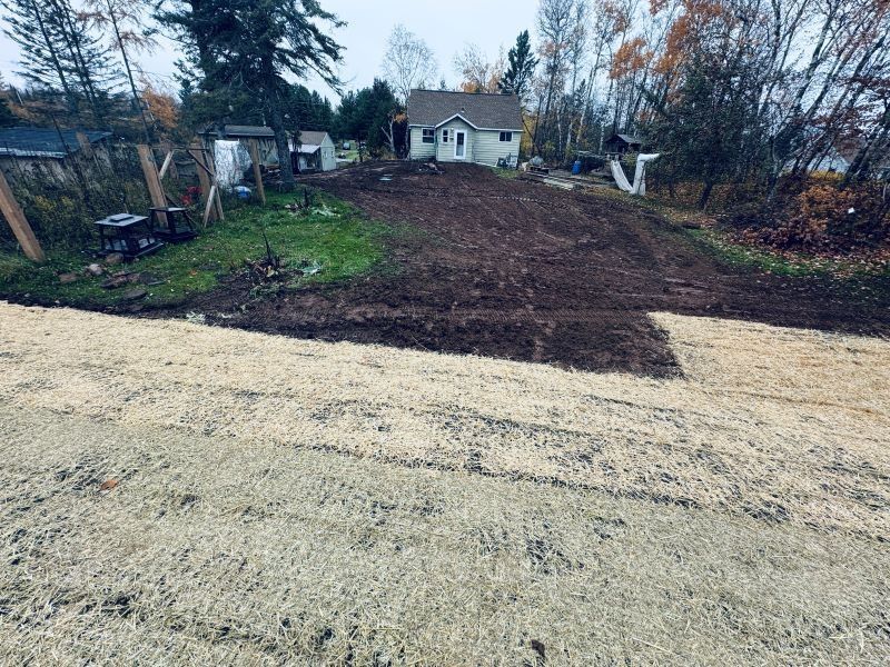 Dirt driveway leads uphill to a small white house, with brown dirt patch in between. Cloudy sky.