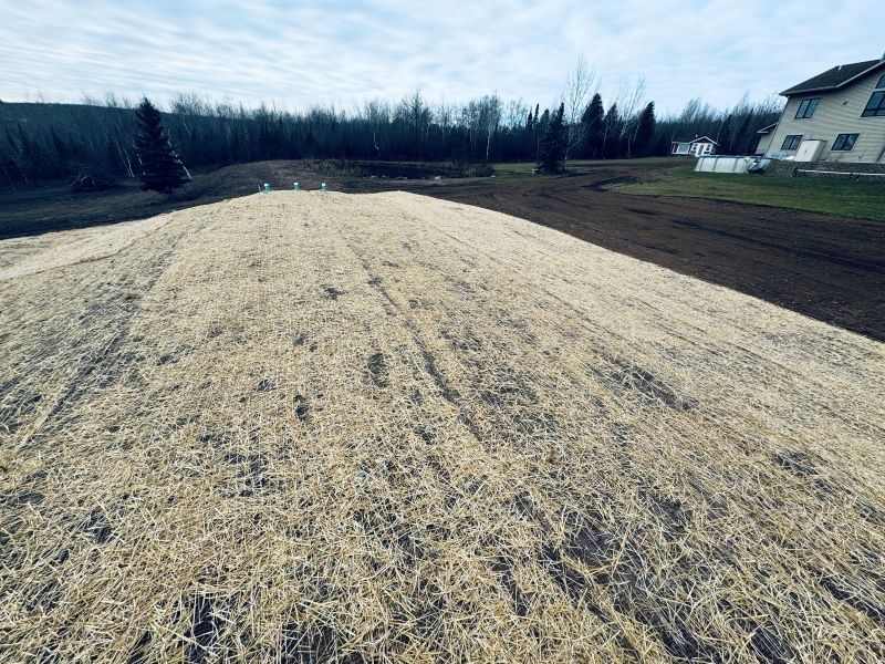 Large brown field covered with straw, some bare brown dirt, house in the background.