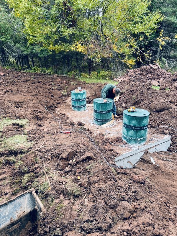 Construction site: worker near three green septic tanks in a dirt field, surrounded by soil, with trees in the background.