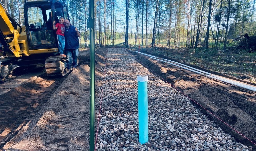 Construction site with gravel-filled trenches, blue pipe, people on a digger, and trees in the background.