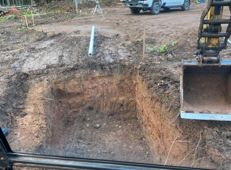 Excavation pit, revealing soil layers.  Backhoe bucket to the right. White truck, survey equipment in the background.