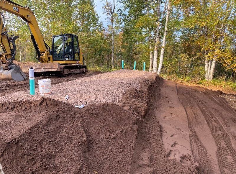 Construction site with a small excavator, gravel, and exposed soil; trees in the background.