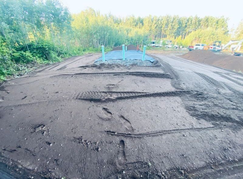 Dirt and gravel construction site with green pipes, trees, and heavy machinery.