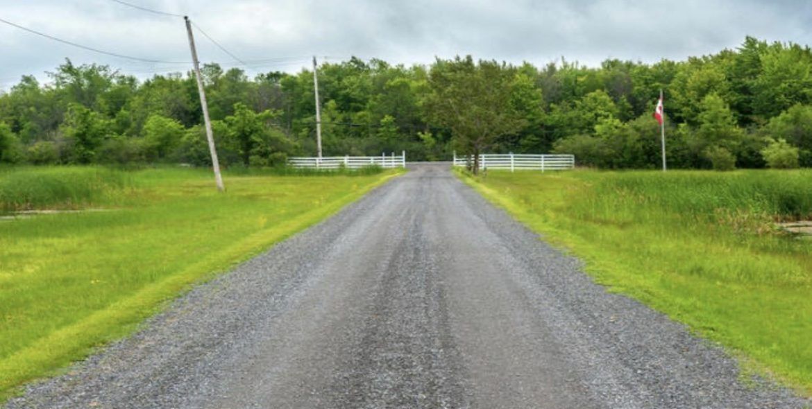 Gravel road leading to a white fence and trees, with a flag on the right and green grass on either side.