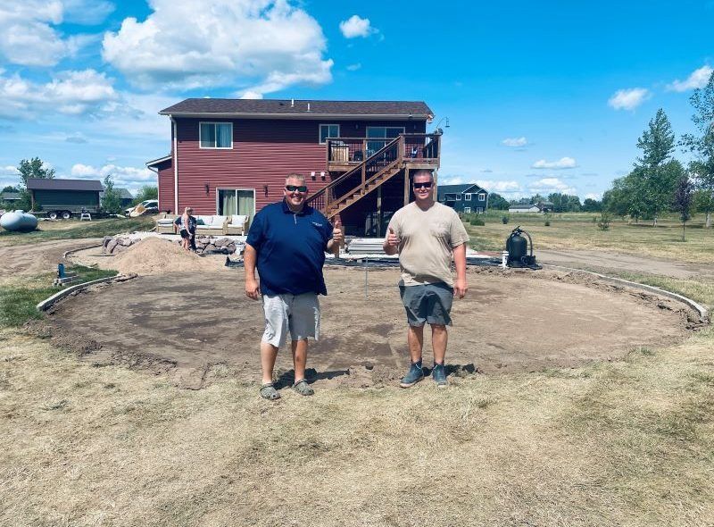 Two men stand outside a house; a circular patio is being built.