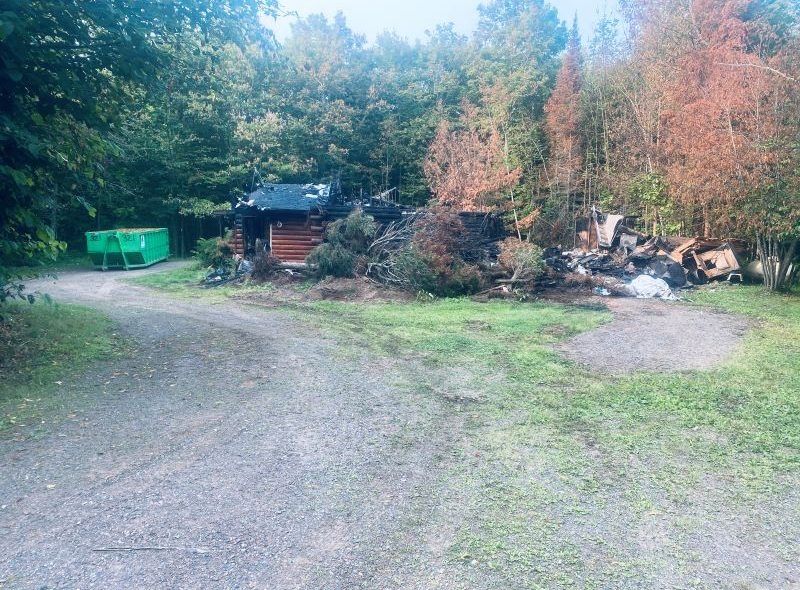 Gravel road leads to a log cabin with a green dumpster, surrounded by trees and brush.
