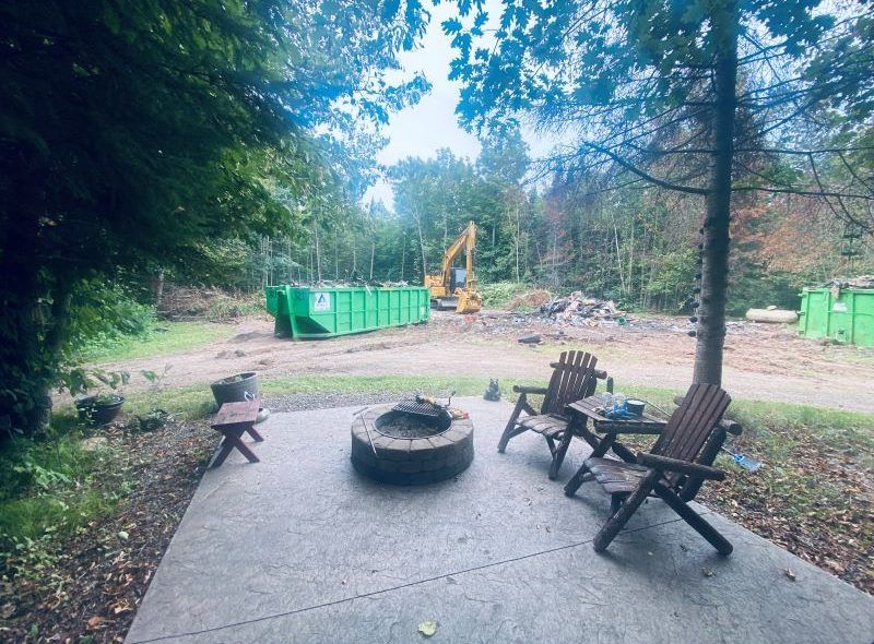 Concrete patio with chairs and fire pit; construction equipment and dumpsters in background.