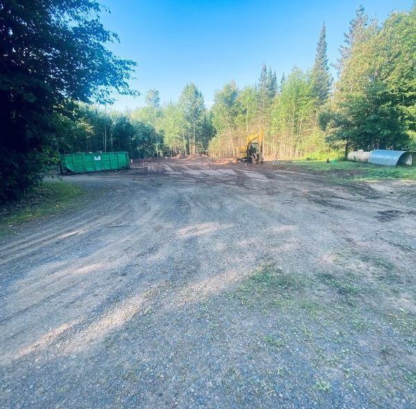 Gravel clearing with trees and green dumpster on either side. An excavator works in the distance on a sunny day.