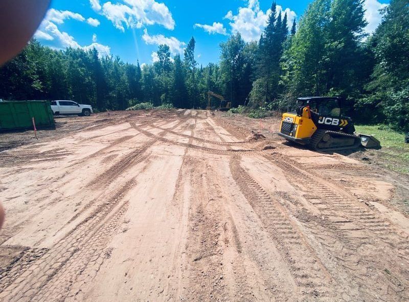 A leveled sandy construction site with a JCB skid steer; trees and a truck are in the background.