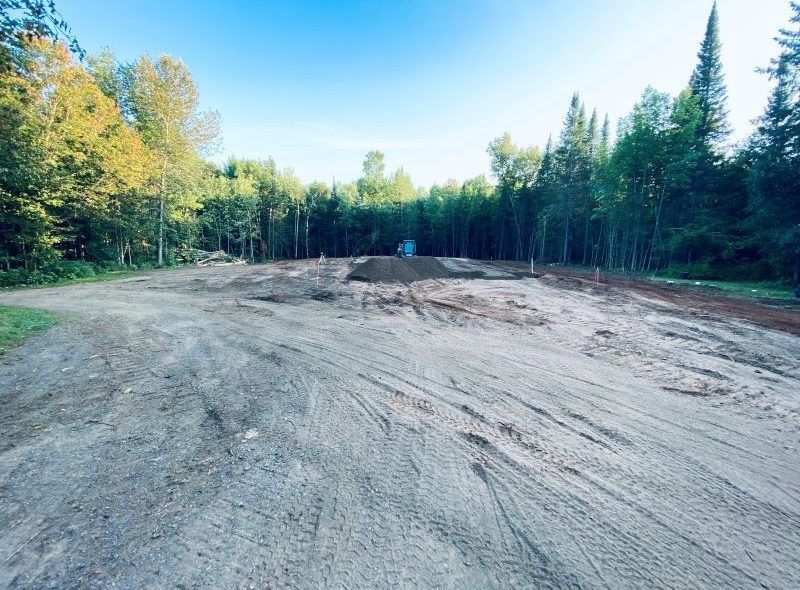 Cleared dirt lot with tire tracks, surrounded by trees, under a blue sky.