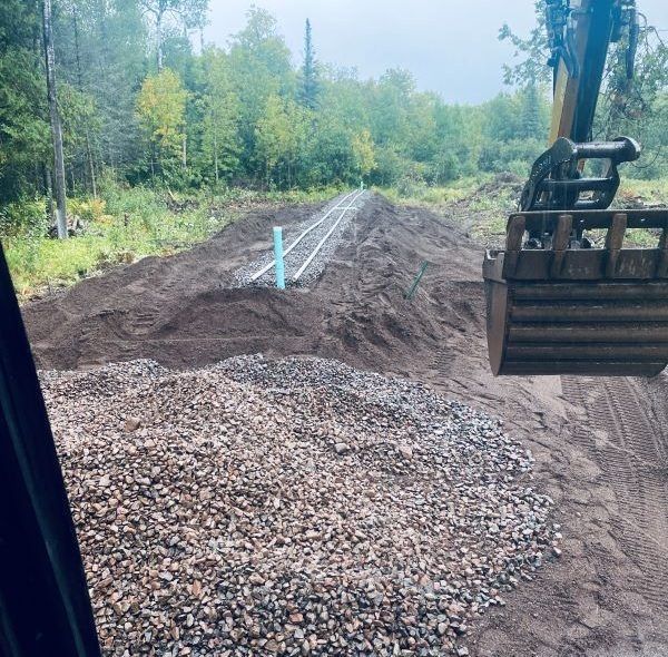 Excavator near a mound of dirt and gravel, with buried pipes, in a wooded area.