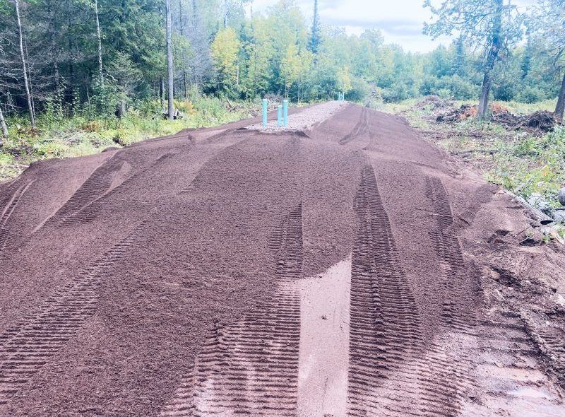Large pile of brown dirt, with tire tracks. Blue pipes stick out, surrounded by trees.