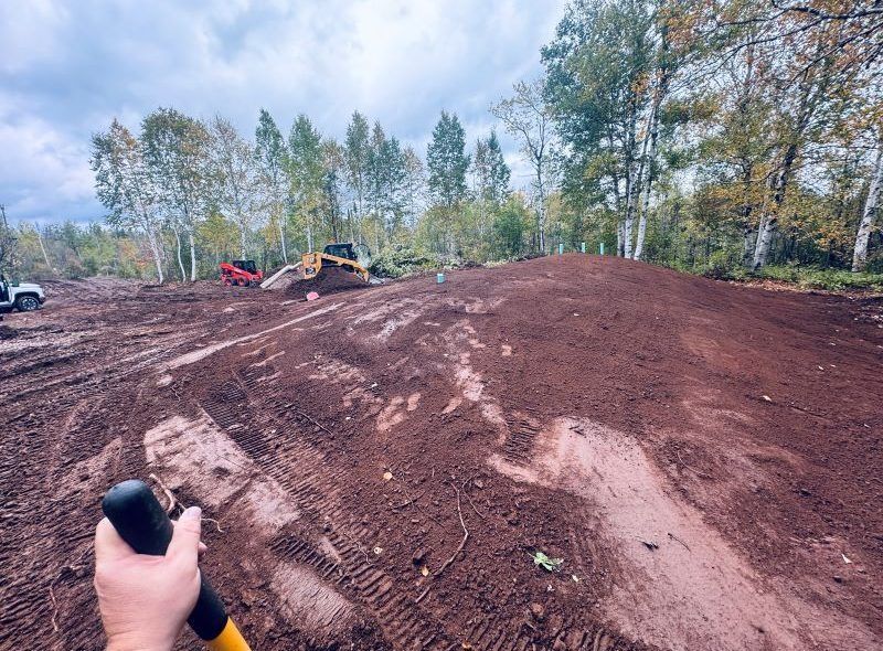 Person holding shovel looking at a dirt mound in a construction site with heavy machinery and trees in the background.
