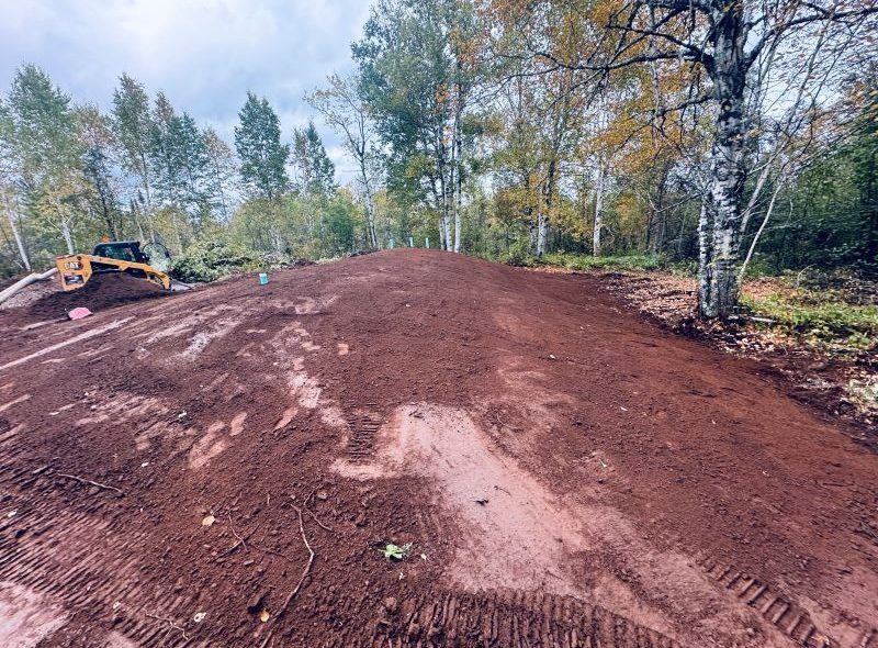 A bulldozer shaping a large mound of red dirt in a wooded area. Overcast sky.