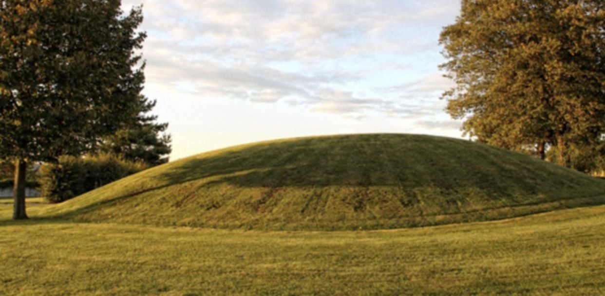 A large grassy mound in a park setting with trees and a cloudy sky in the background.