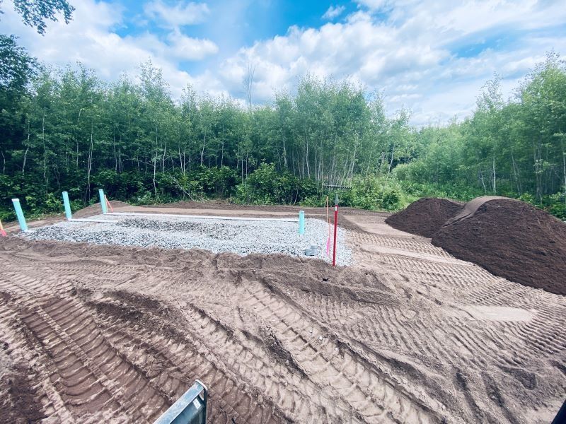 Construction site with gravel foundation, blue pipes, dirt piles, and trees under a cloudy sky.