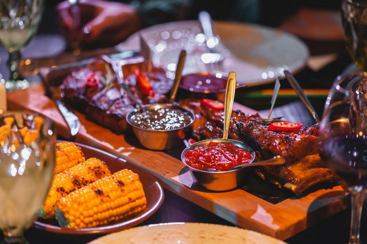 Grilled meats and corn on a wooden table with sauces and drinks, ready for a meal
