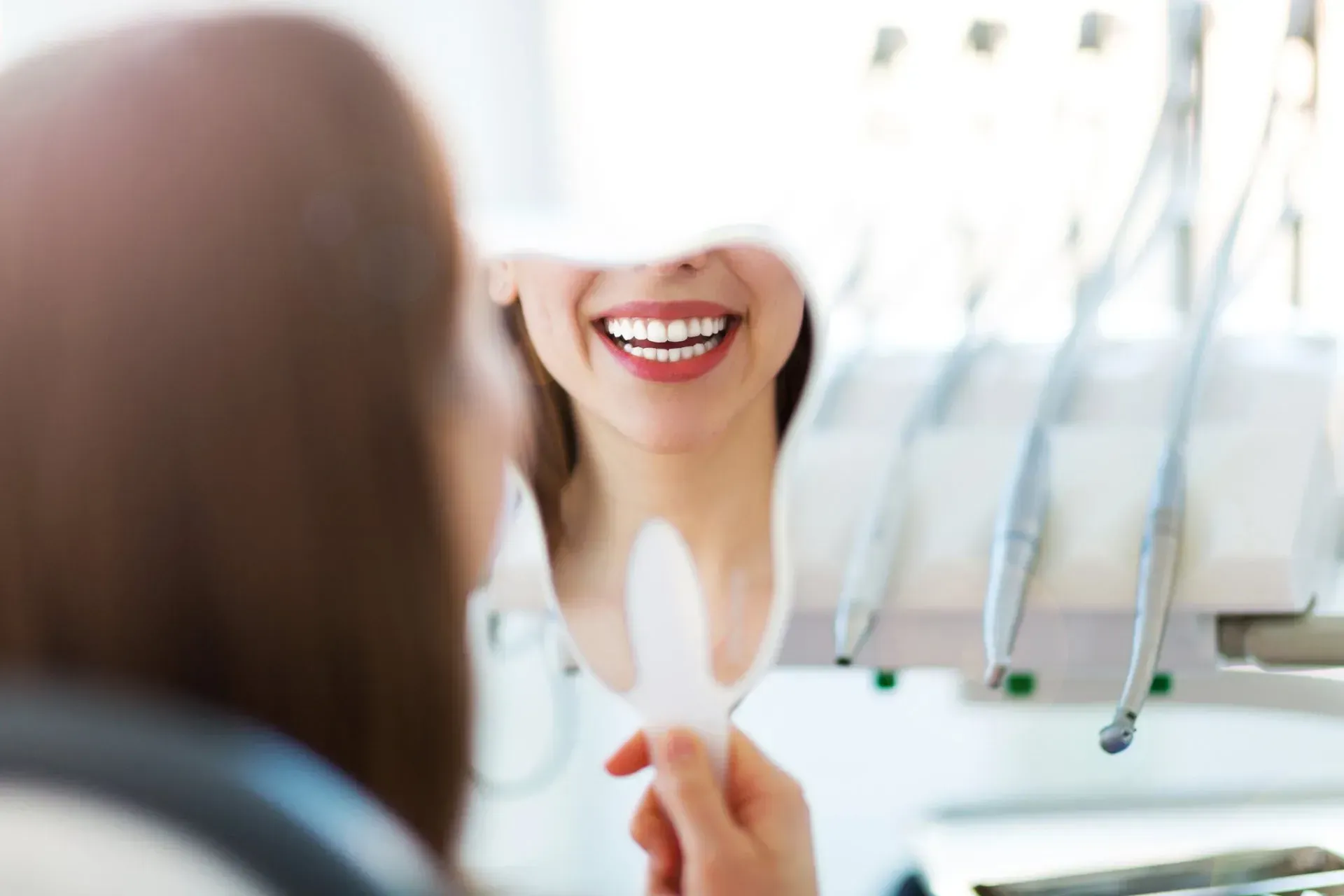 Una mujer sonriente sentada en el sillón del dentista se mira los dientes en un espejo con los instrumentos dentales cerca.