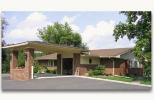 A single-story brick building with a covered drive-through entrance, surrounded by trees under a blue, cloudy sky.