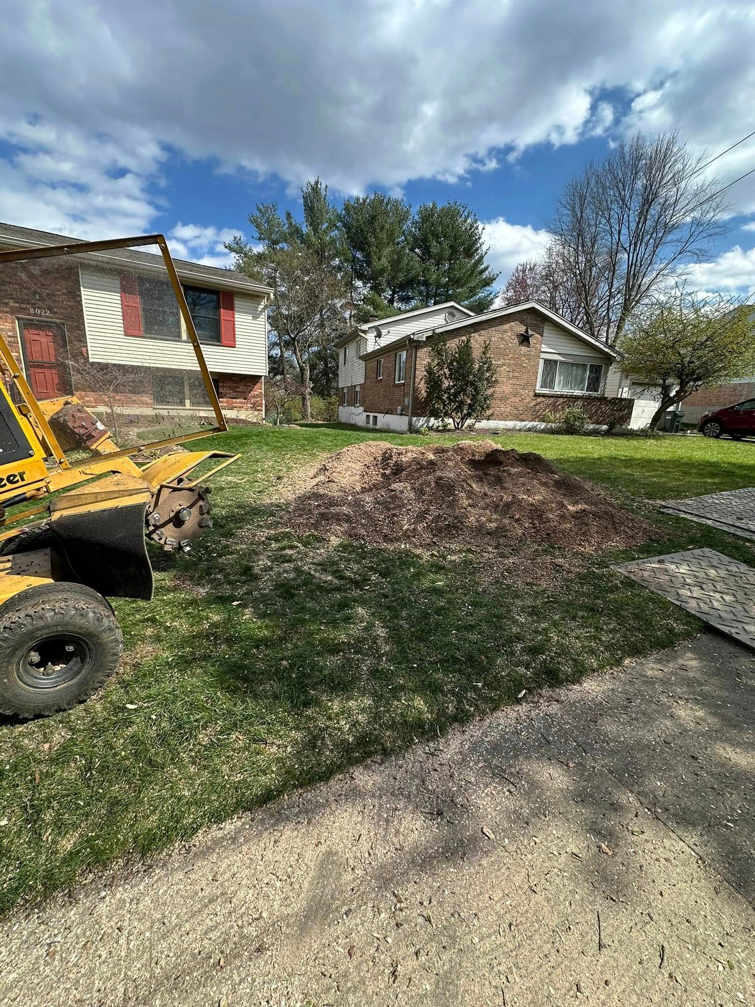 Front yard with a mulch pile, small houses, blue sky, and part of a yellow skid steer.