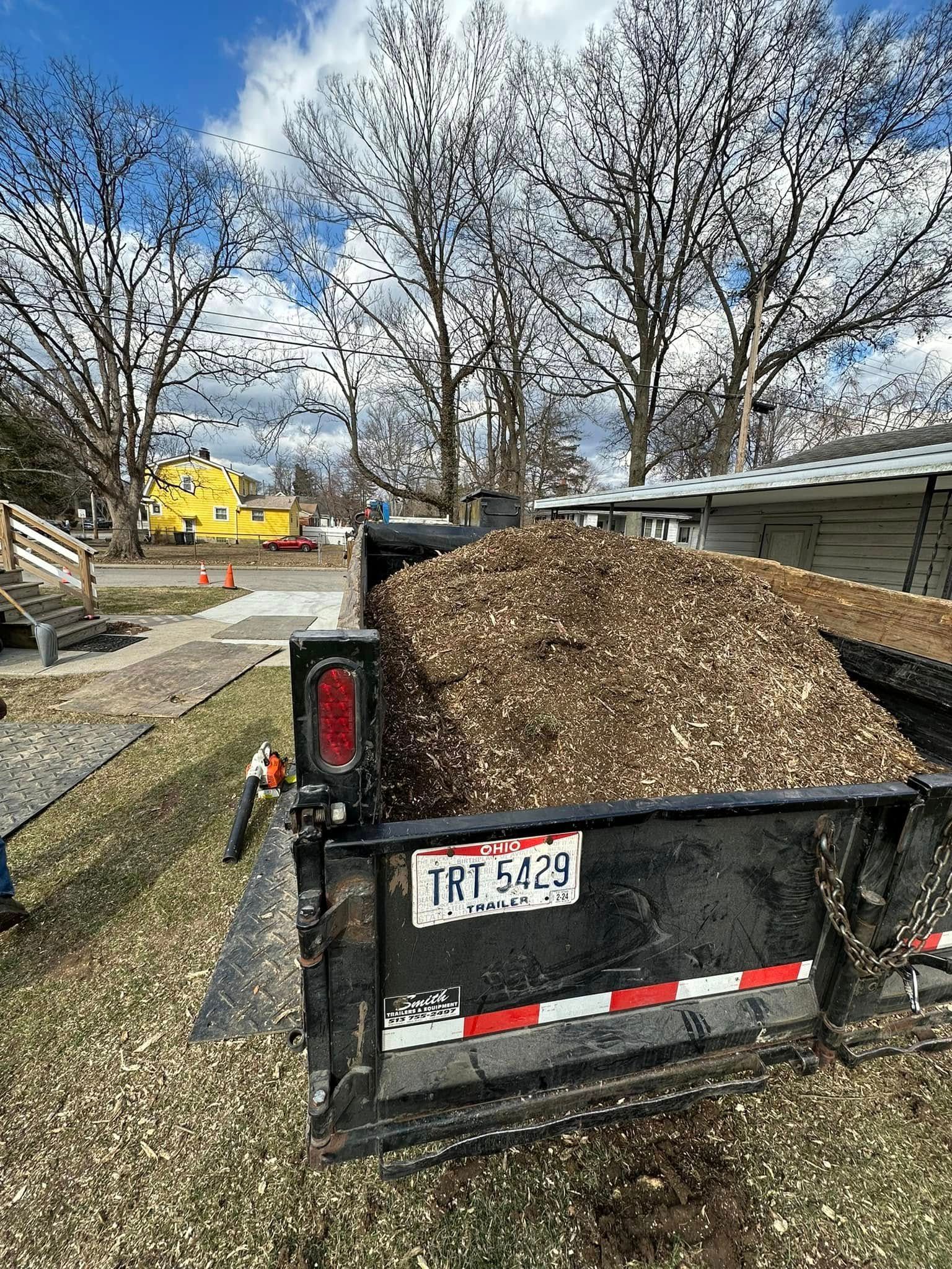 Black utility trailer piled with wood chips beside a gravel path and leafless trees