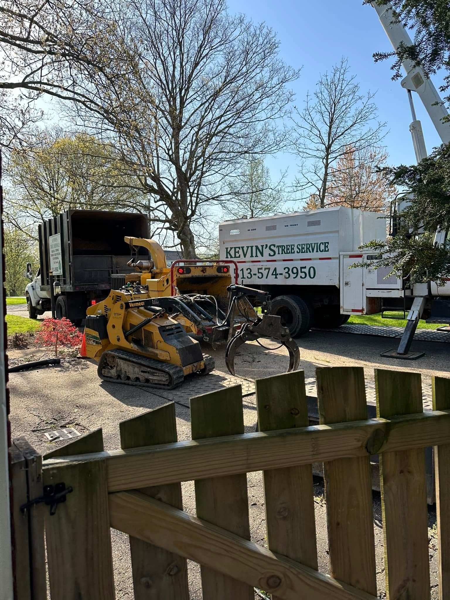Yellow wood chipper by open truck in a yard, with trees and a fence in the foreground.