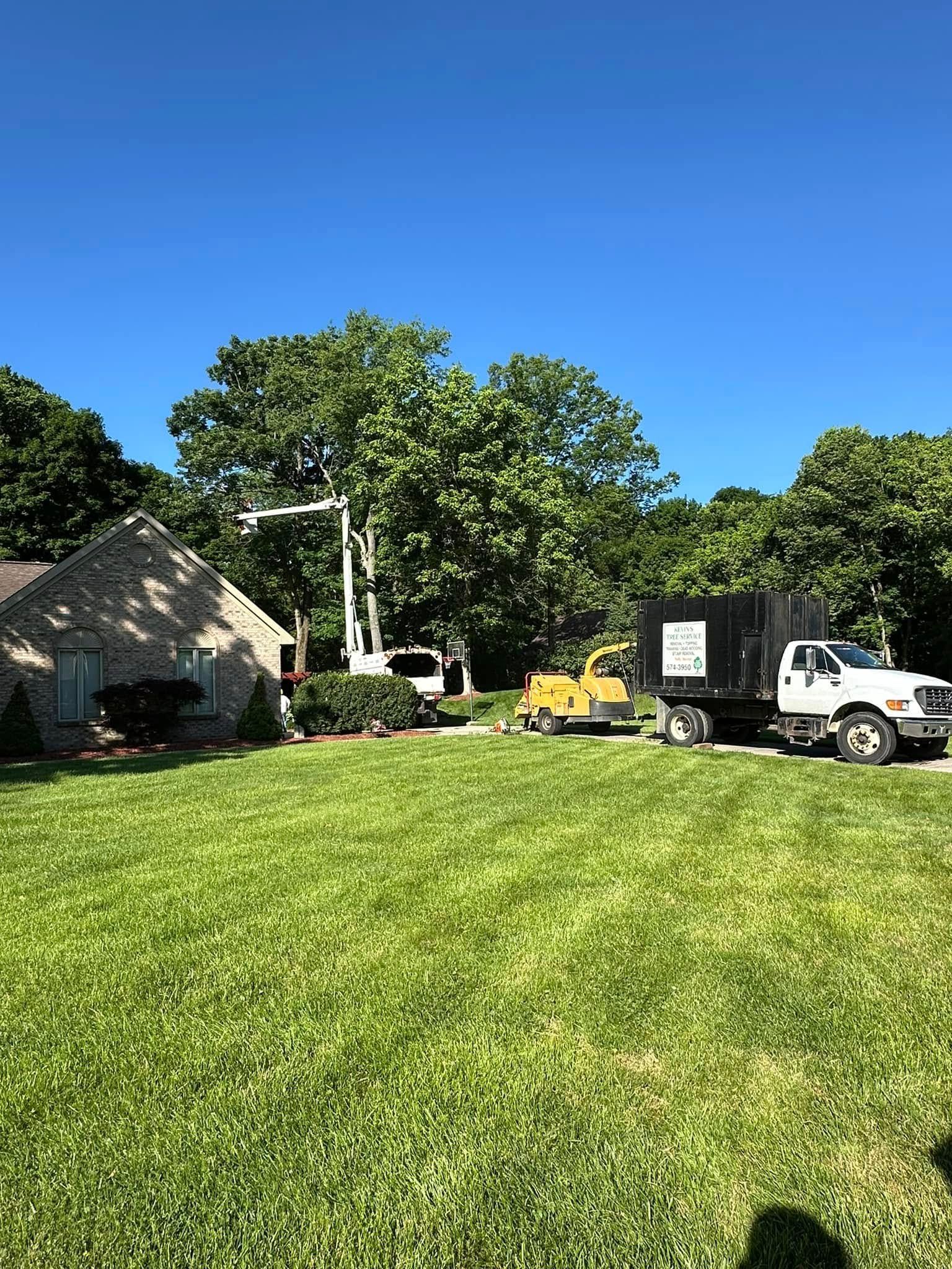 Grassy yard beside a stone house, with a white truck, yellow equipment, and trees under a clear blue sky