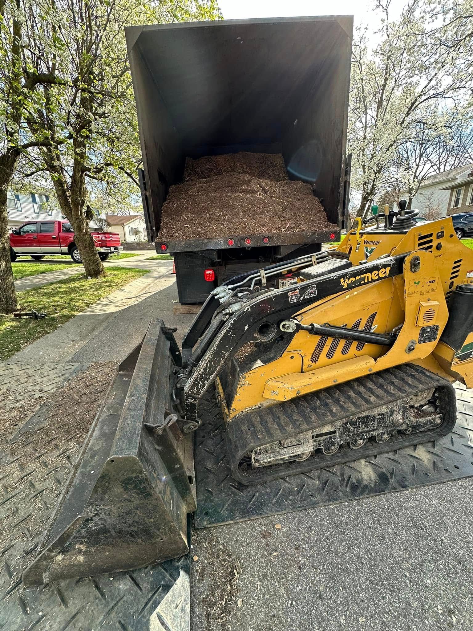 Yellow skid-steer loader dumping gravel into a black trailer on a tree-lined street