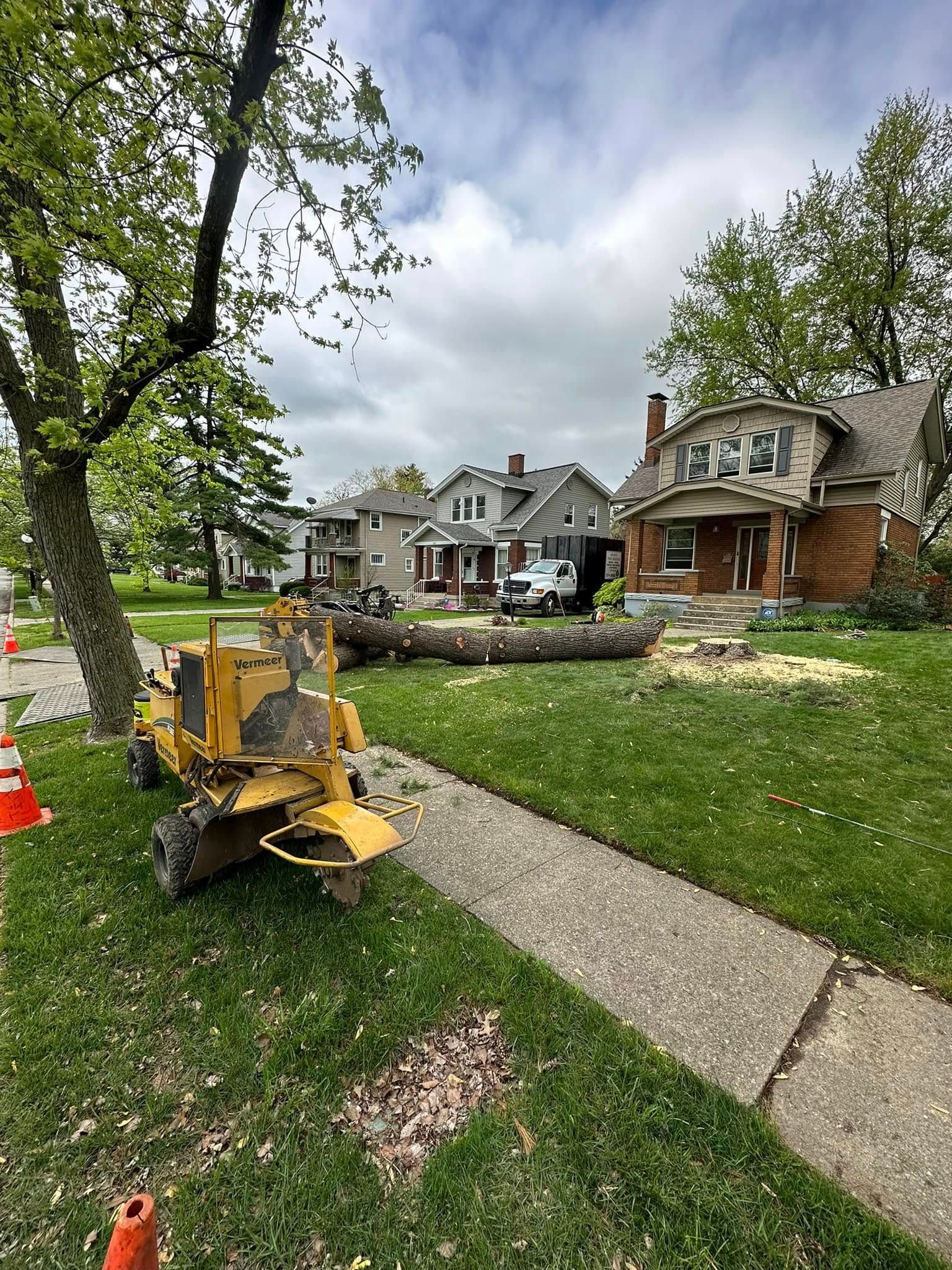 Yellow utility machine on a sidewalk beside a suburban lawn and houses, with orange cones nearby
