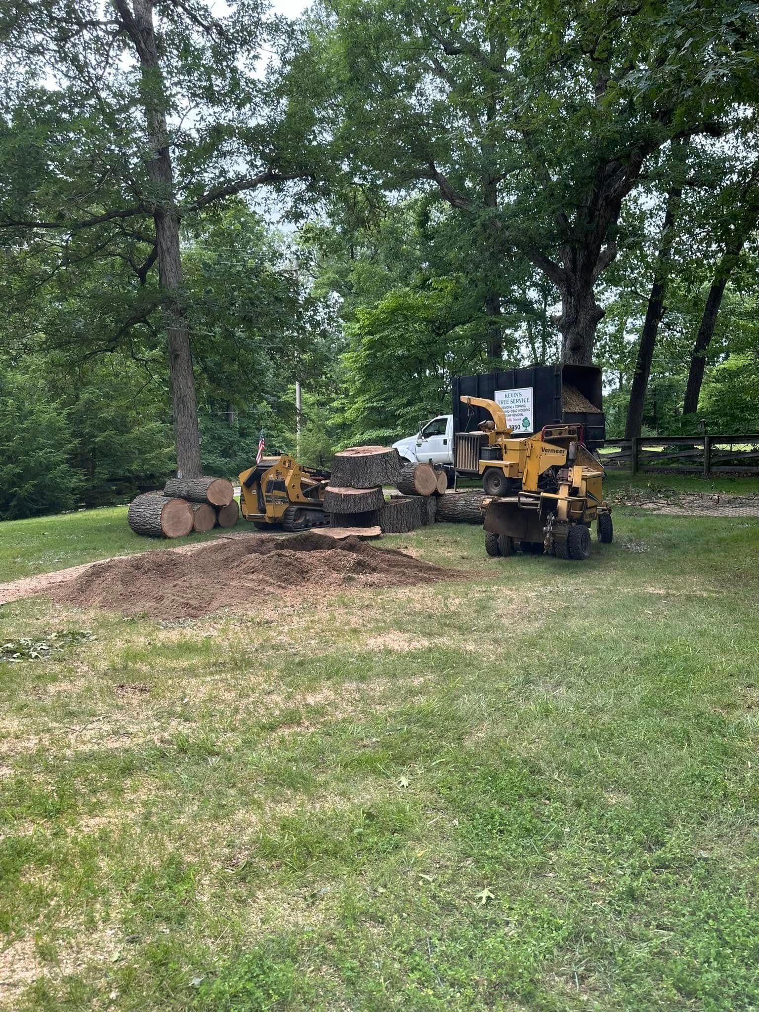 Yellow excavator clearing dirt near fallen trees in a grassy wooded area.