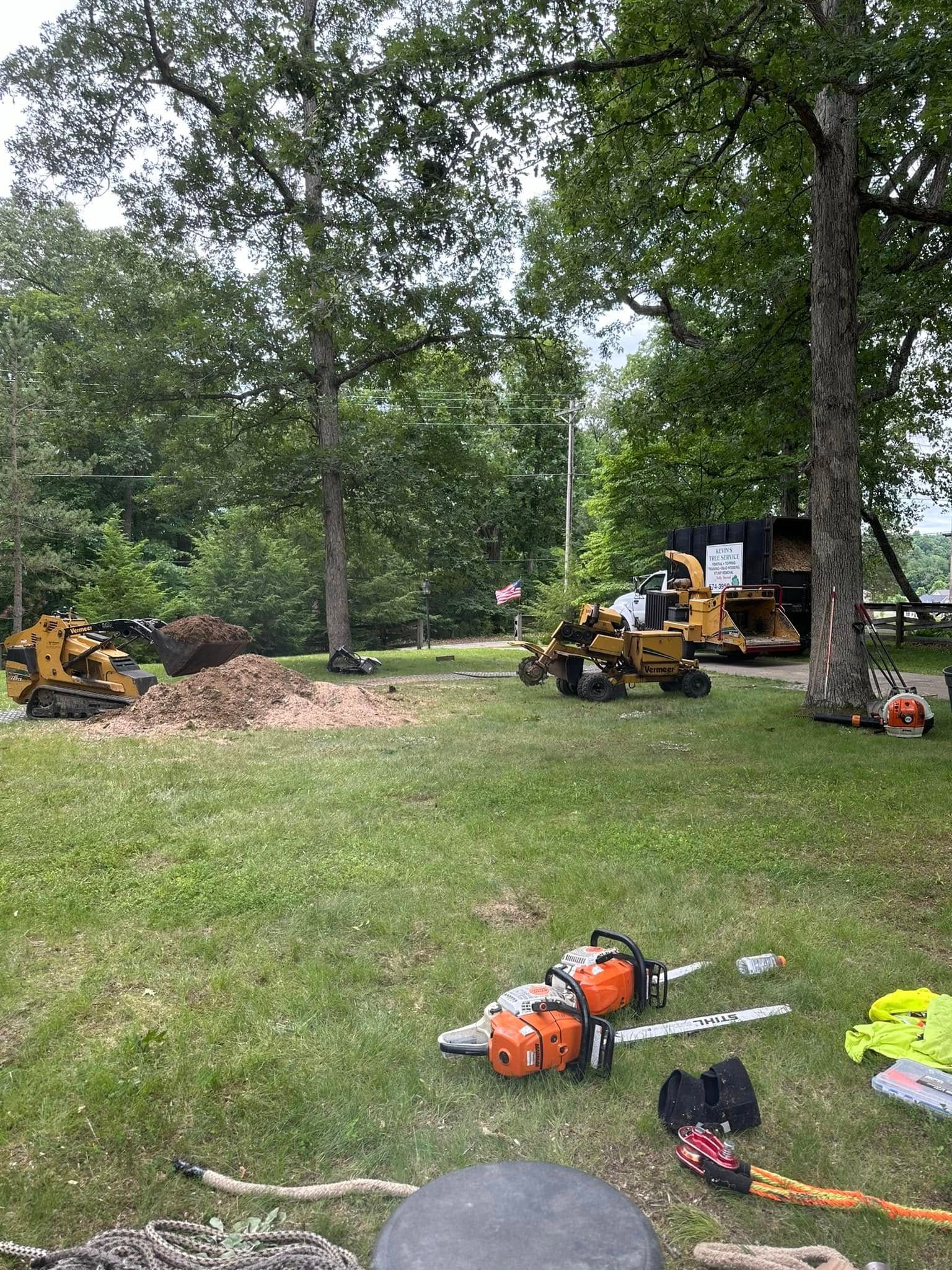 Tree clearing site with a yellow excavator, logs, and landscaping tools in a grassy yard