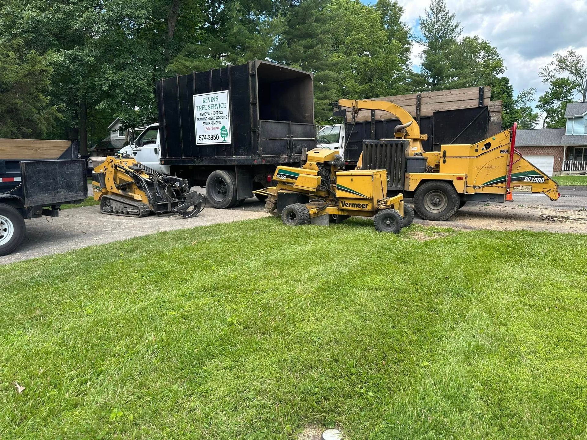 Yellow wood chipper and dump truck in a grassy yard beside a gravel drive, with trees in the background