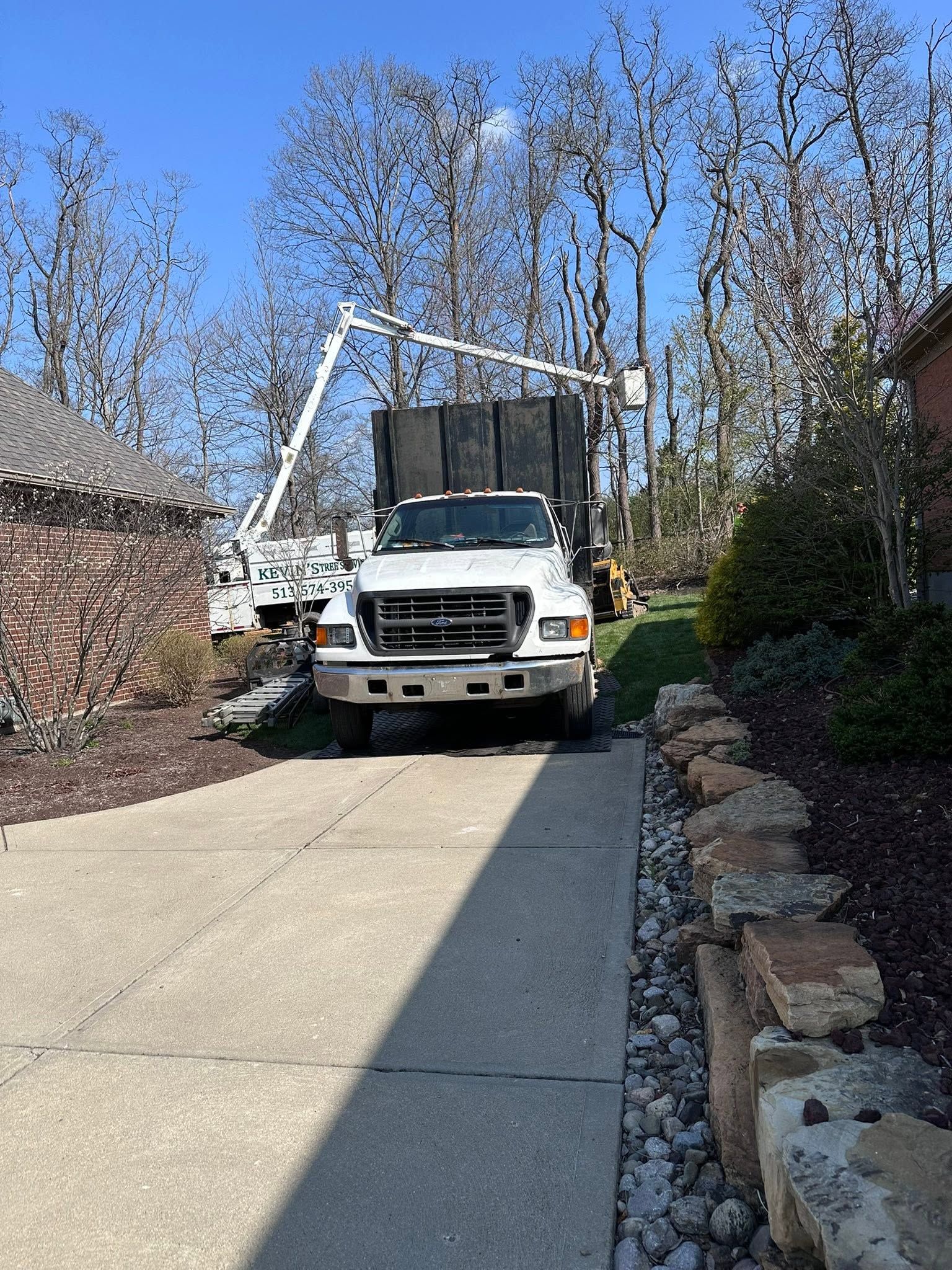 White utility truck parked on a narrow driveway beside stone landscaping and leafless trees on a sunny day