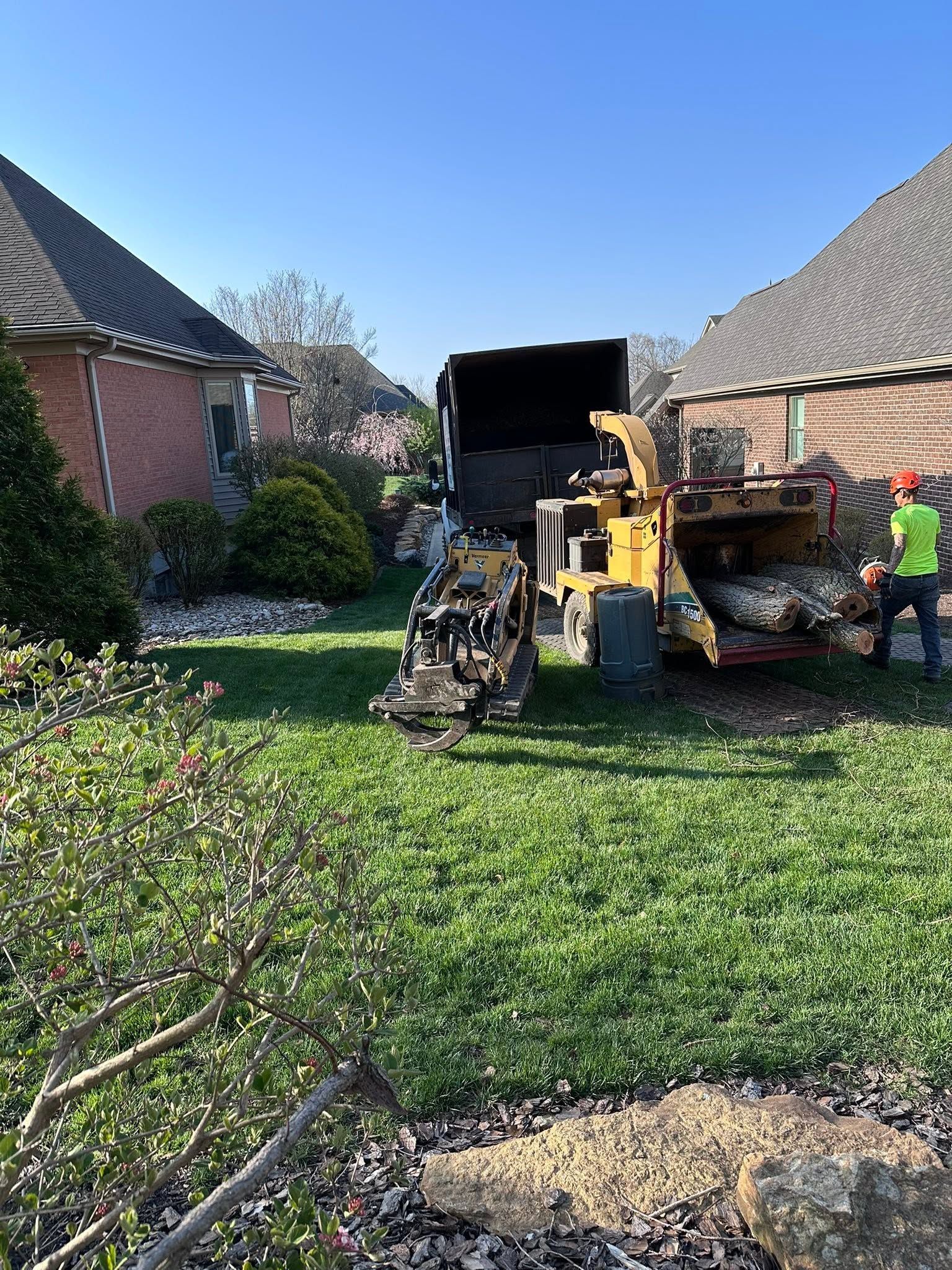 Construction equipment in a grassy residential yard, with a worker beside a dump truck between two houses.