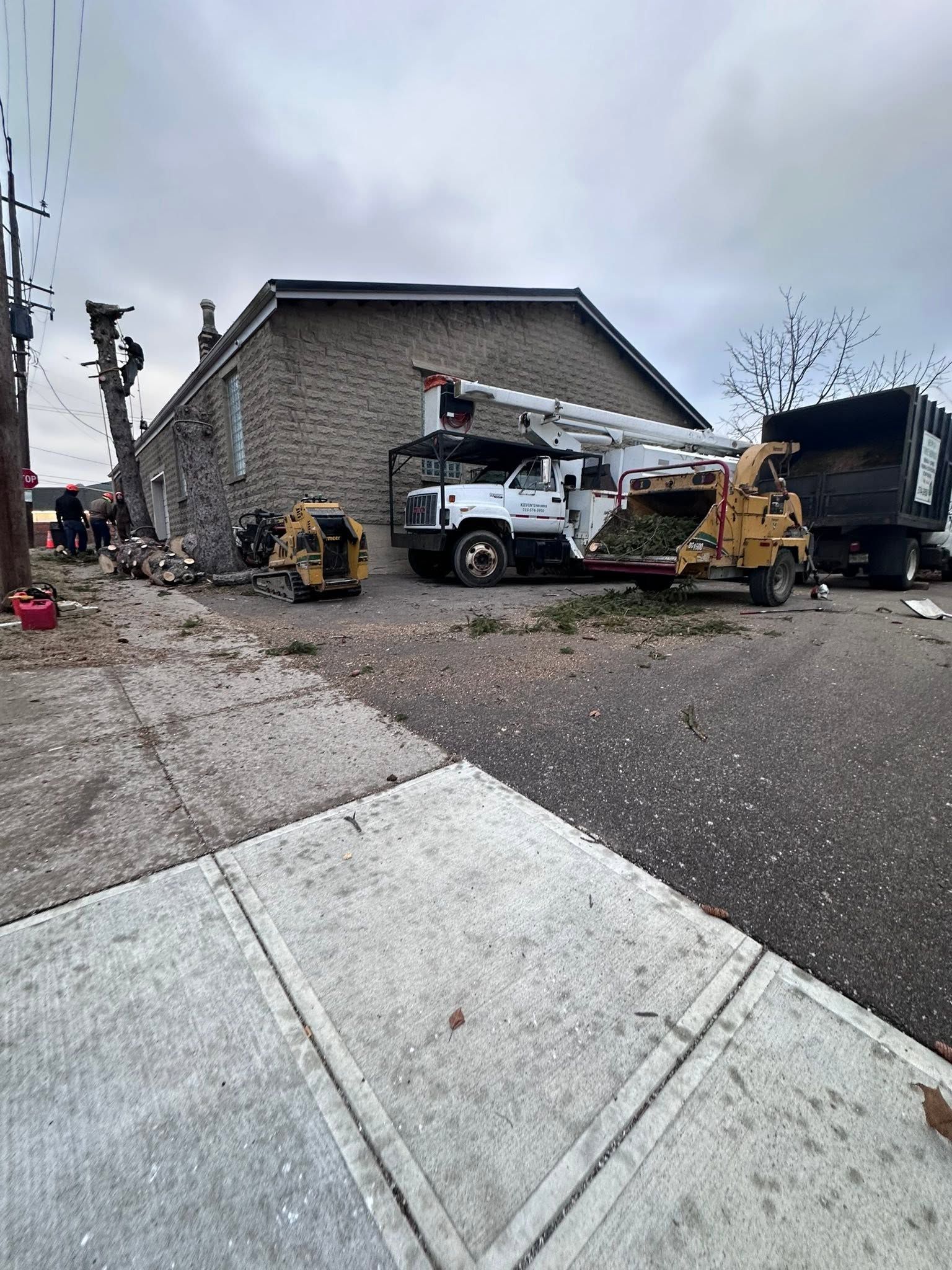 Construction truck parked beside a stone house with a yellow excavator and utility workers on a driveway