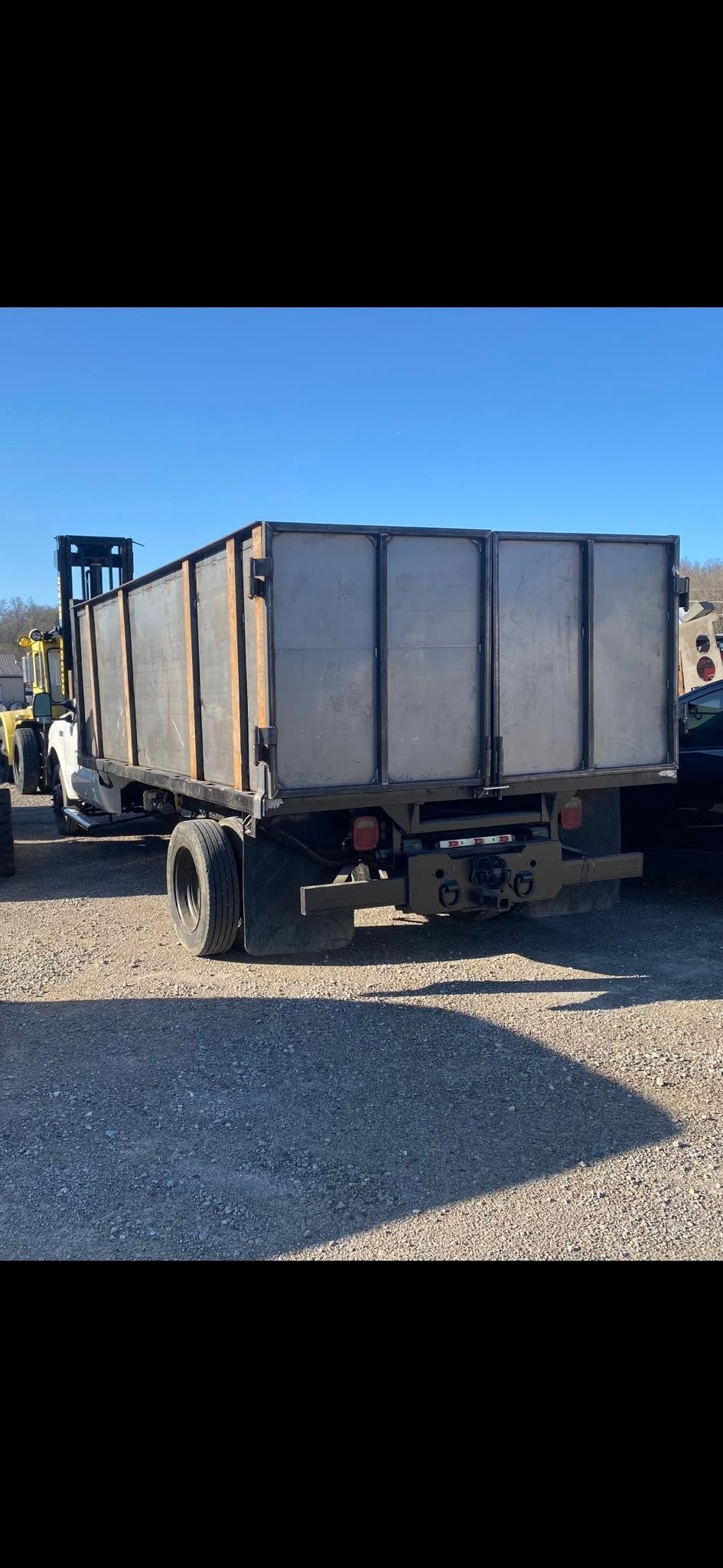 Rear view of a gray dump trailer parked on gravel under a clear blue sky.
