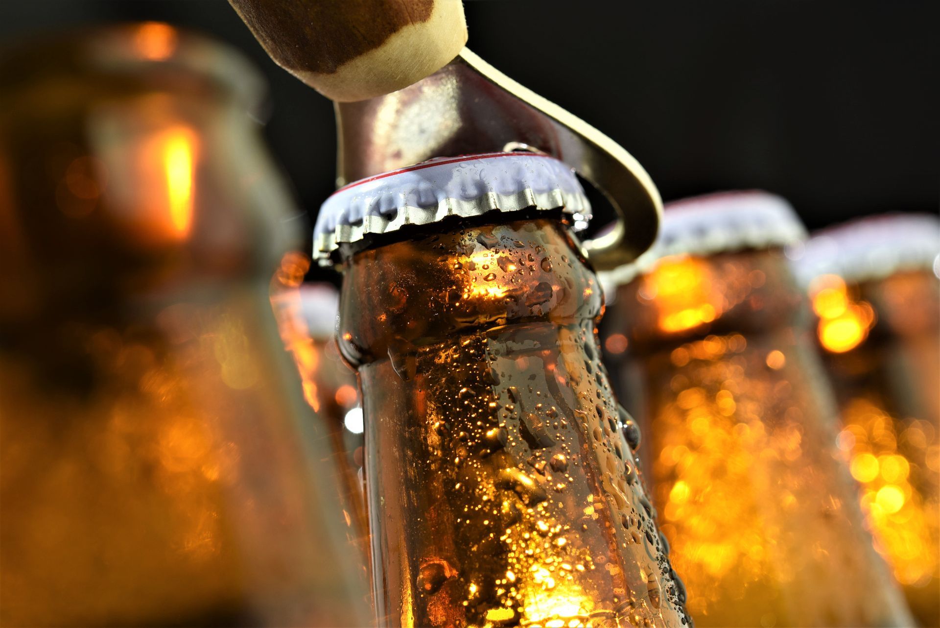 Un apribottiglie che rimuove il tappo da una bottiglia di birra marrone e bagnata, con altre bottiglie sullo sfondo.