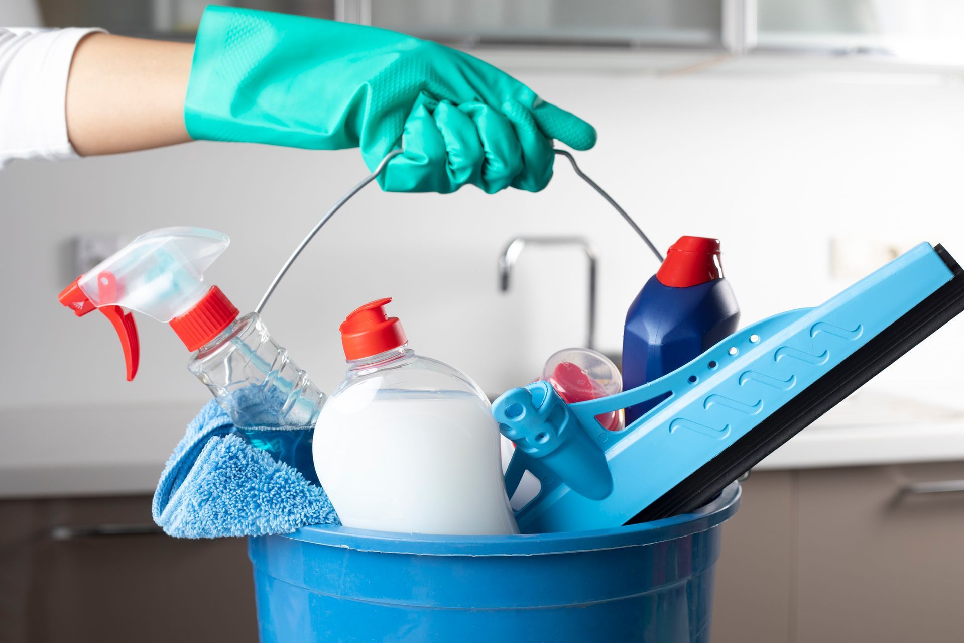 Persona che tiene in mano un secchio blu con prodotti per la pulizia, tra cui flaconi spray, detergente e un tergivetro.