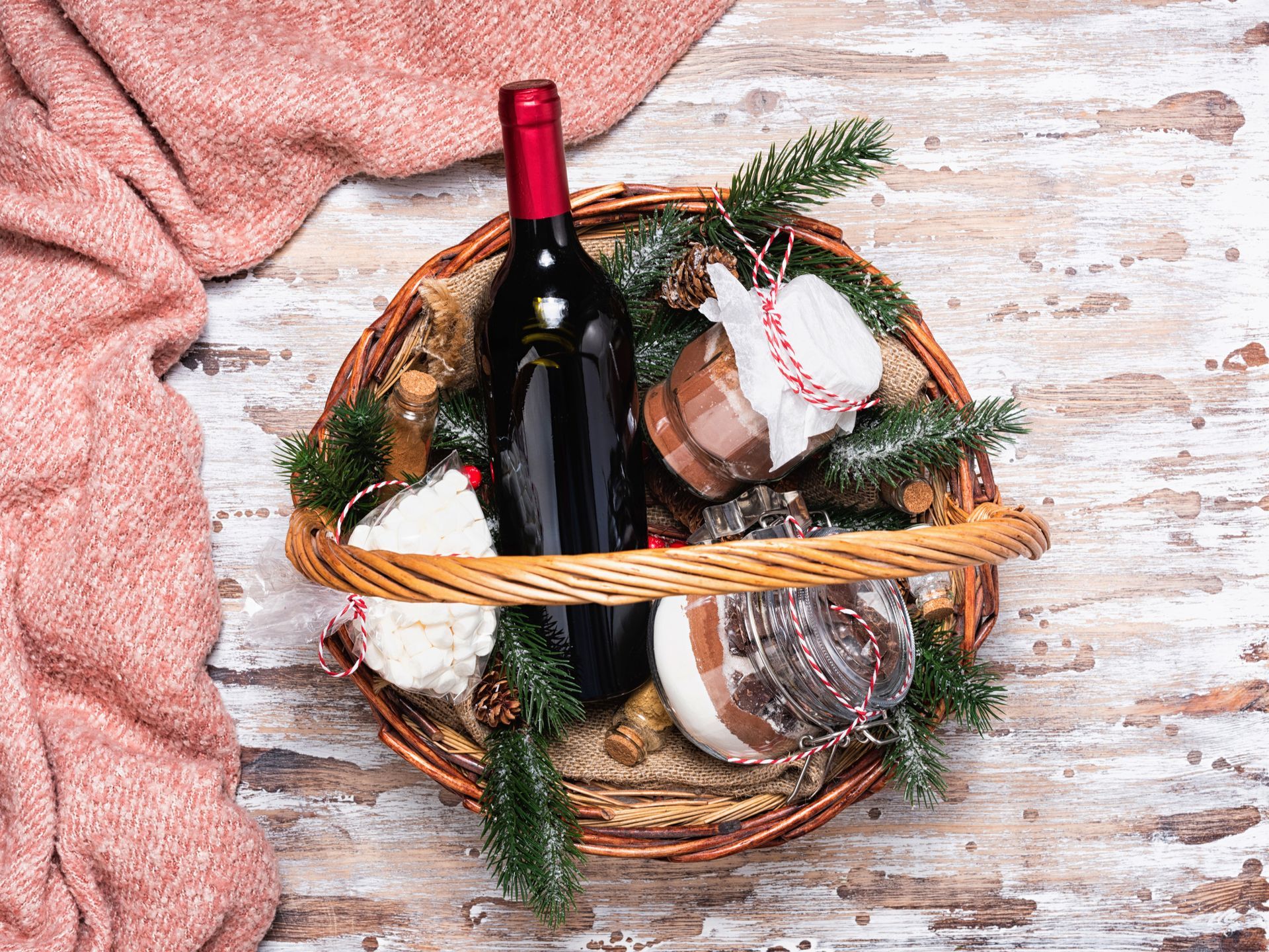 Set regalo di cesti di vimini con vino rosso, barattoli e rami di pino, su una superficie di legno invecchiato e una coperta rosa.