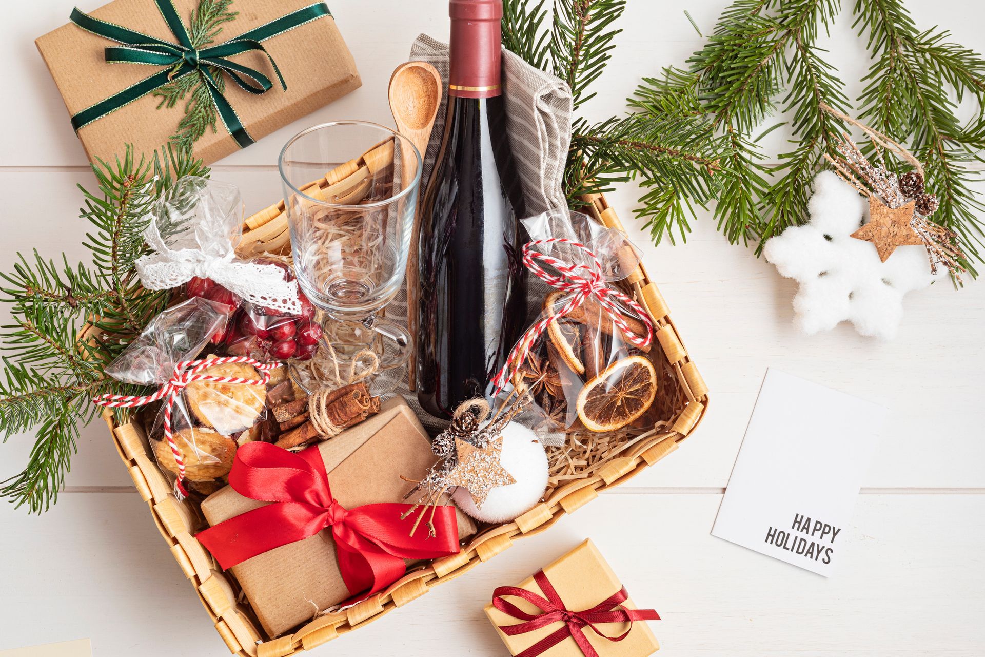 Cesto regalo con vino rosso, bicchieri, regali incartati e decorazioni natalizie.