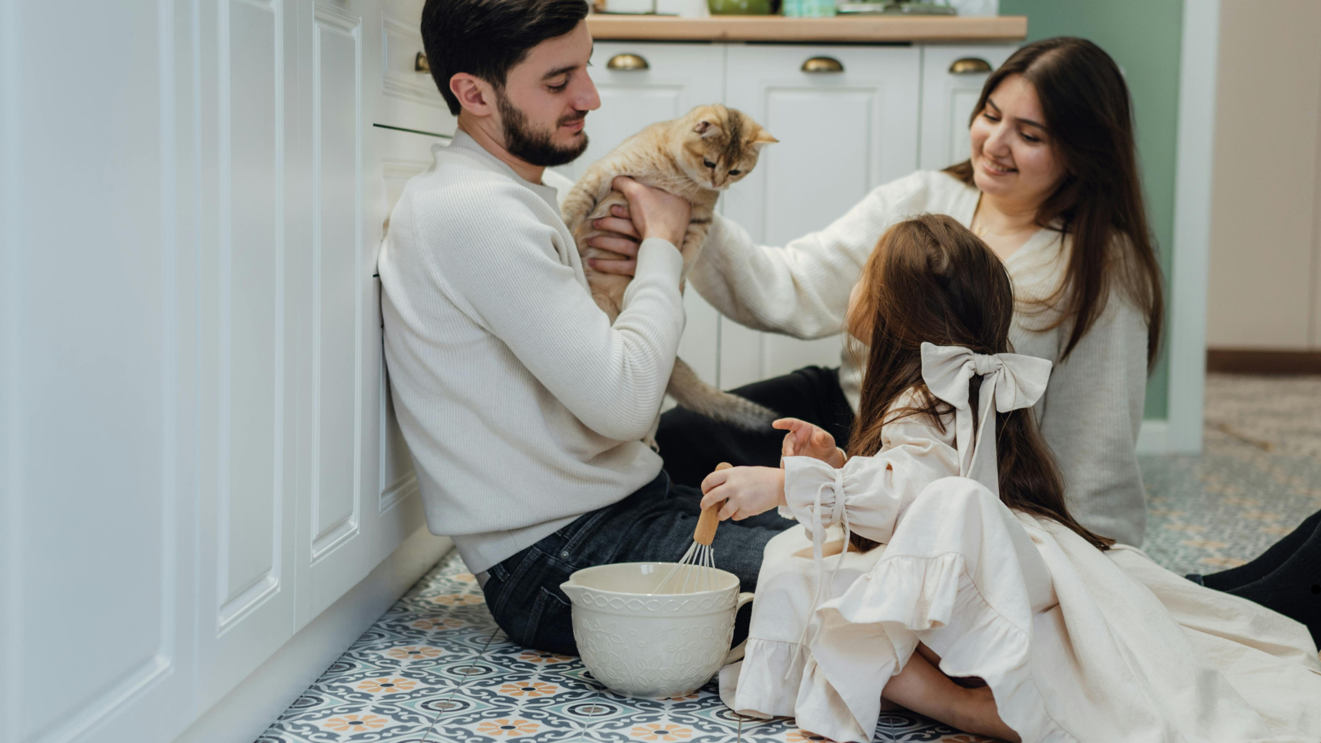 Family of three in a kitchen playing with a cat while a young girl stirs a mixing bowl on the floor, smiling and enjoying quality time together.
