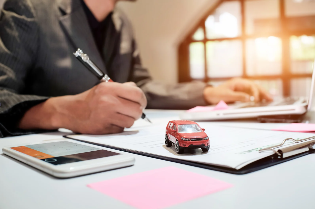 Person writing on paper, red toy car, calculator, and laptop on desk.