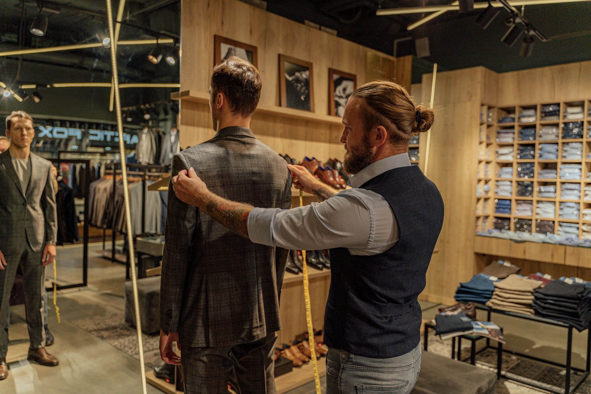 A tailor measures a customer's suit jacket in a clothing store, with shelves of folded garments in the background.