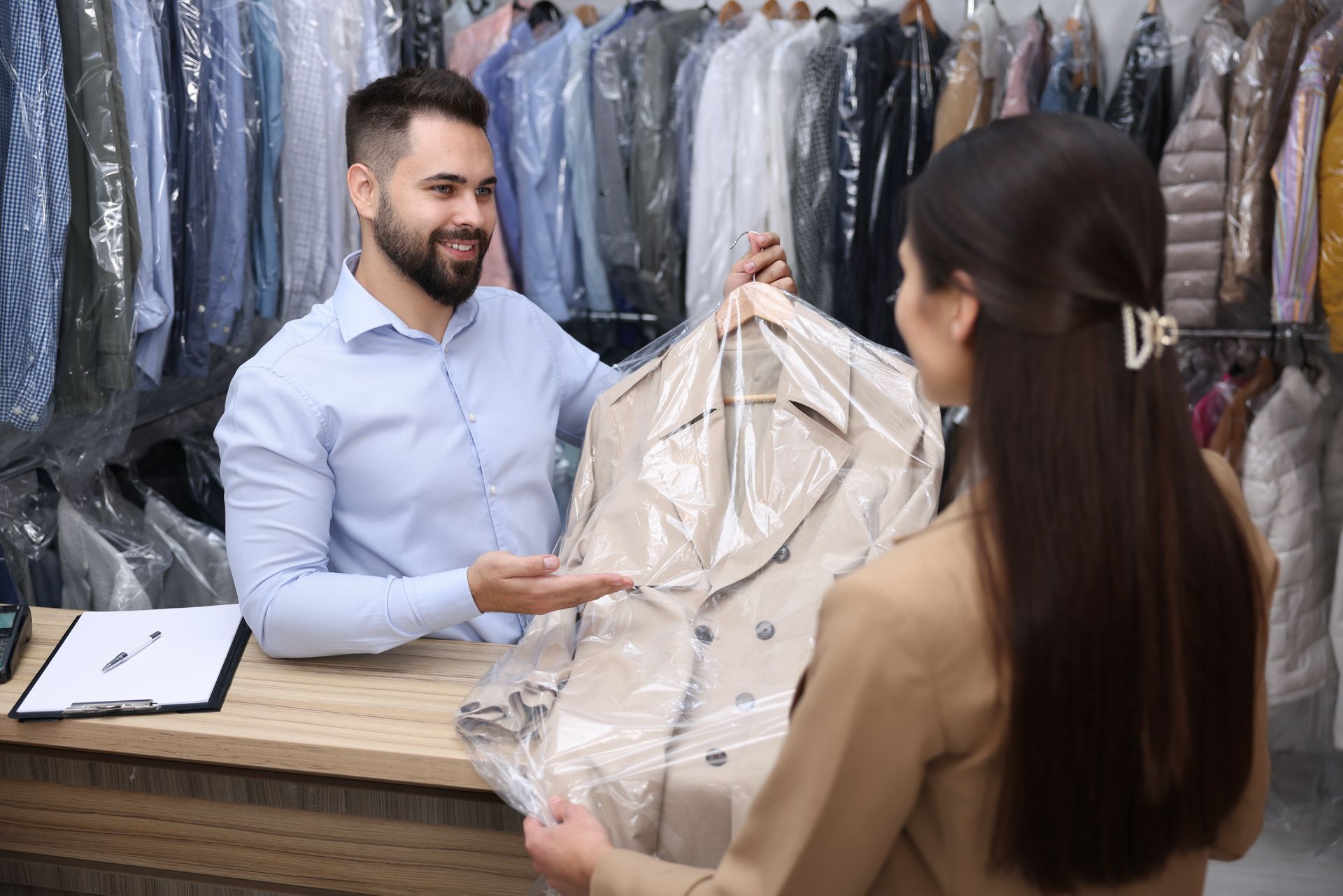 A person at a dry cleaner's counter handing a bagged tan coat to a customer.