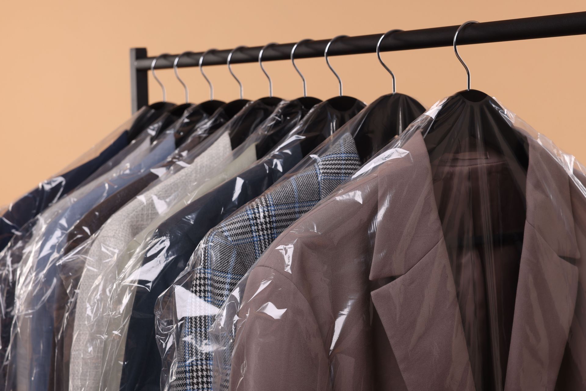 A row of suit jackets in plastic garment bags hanging on a rack against a solid beige background.